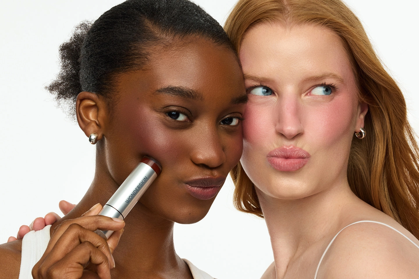 Two women with blush on their cheeks, one holding a blush stick to her face